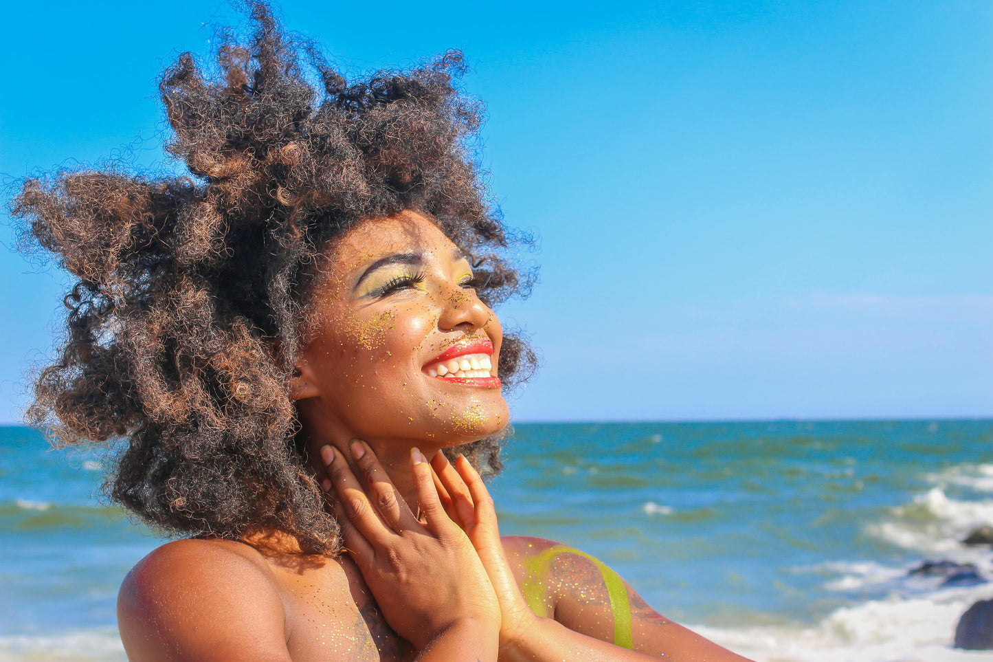 Joyful woman with glitter on her face and body smiling brightly at the beach under a clear blue sky, embracing natural beauty and self-confidence.