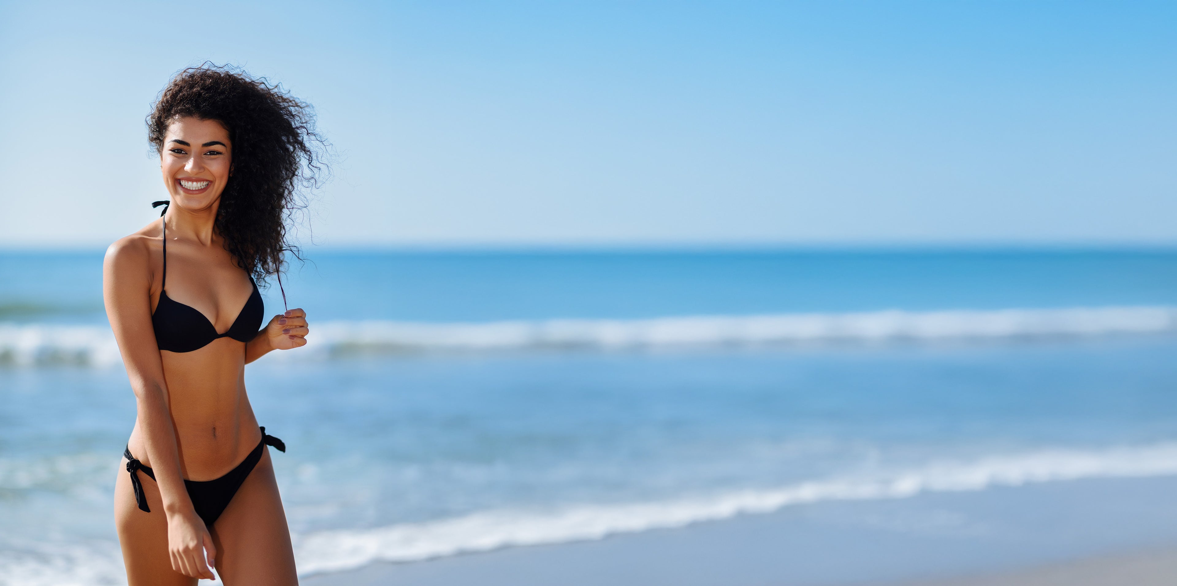 Smiling woman with curly hair blowing in the wind wearing a black bikini with the ocean waves in the background