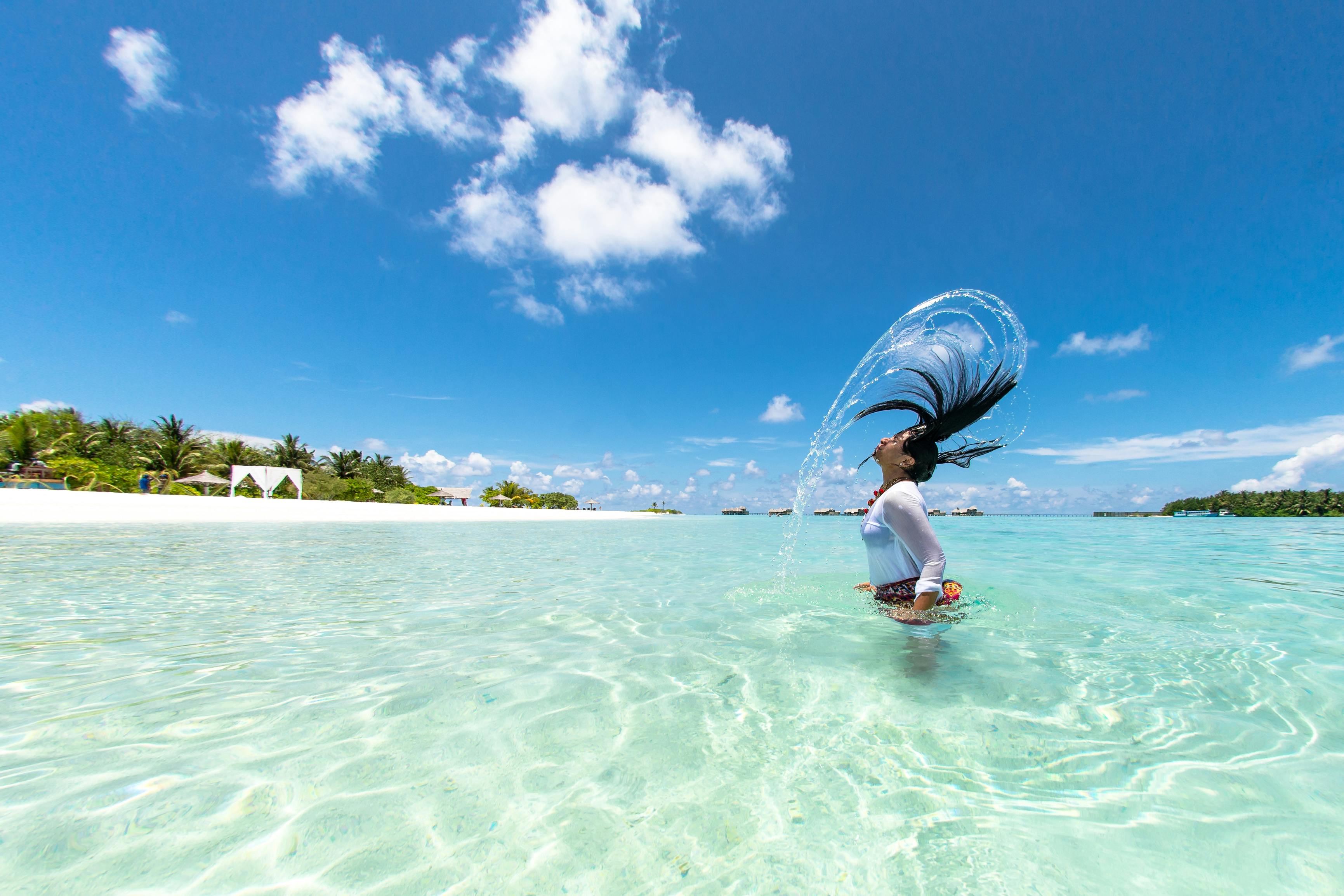 Woman flipping her hair in crystal-clear turquoise water at a tropical beach, creating a water arc under a bright blue sky with scattered clouds.